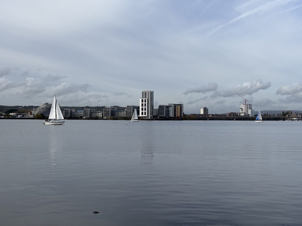A picture over Cardiff bay with a small number of yachts sailing by, which look like the skippers are getting sailing lessons. The boats look about 26 feet long. There is one tall building on the other side of the shore.