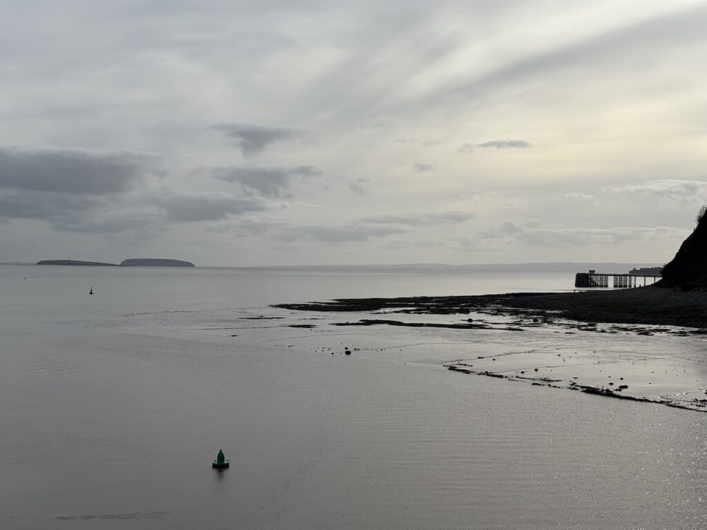 Looking out over the Bristol Channel towards England. The sky is grey, to the left on the horizon are Flat Holm and Steep Holm Islands. There is murky water in the foreground. To the right the start of a cliff edge and just beyond Penarth Pier is visible.