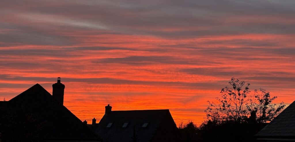 A deep red sky, where the sun is lighting up the underneath of the cloud layer, against a backdrop of suburban houses in silhouette.